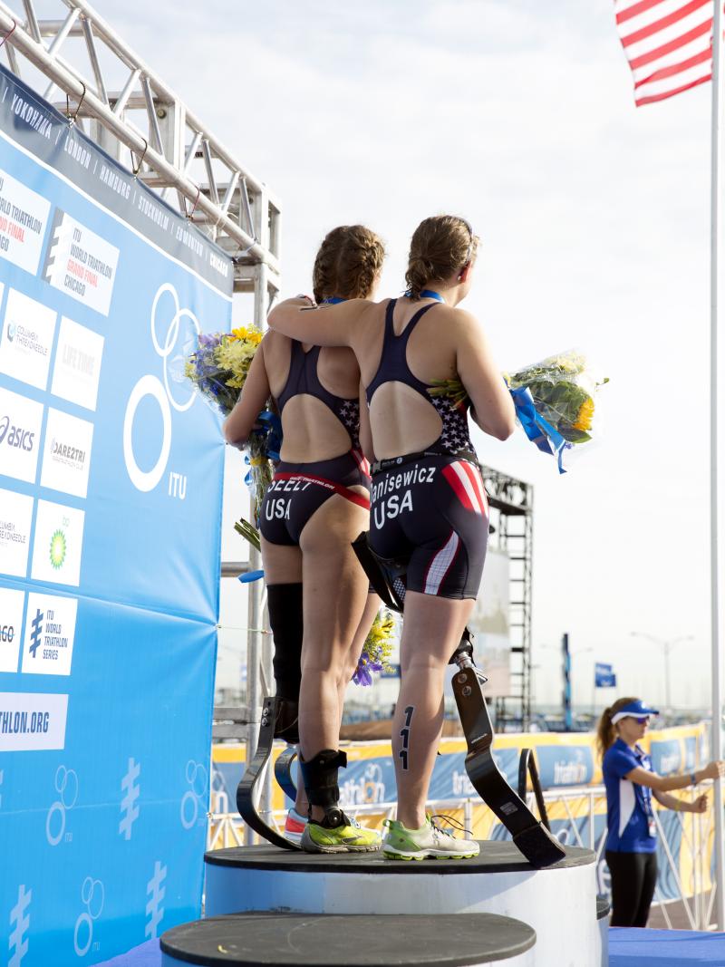 Three Para triathlete women on the podium while the US flag is being raised