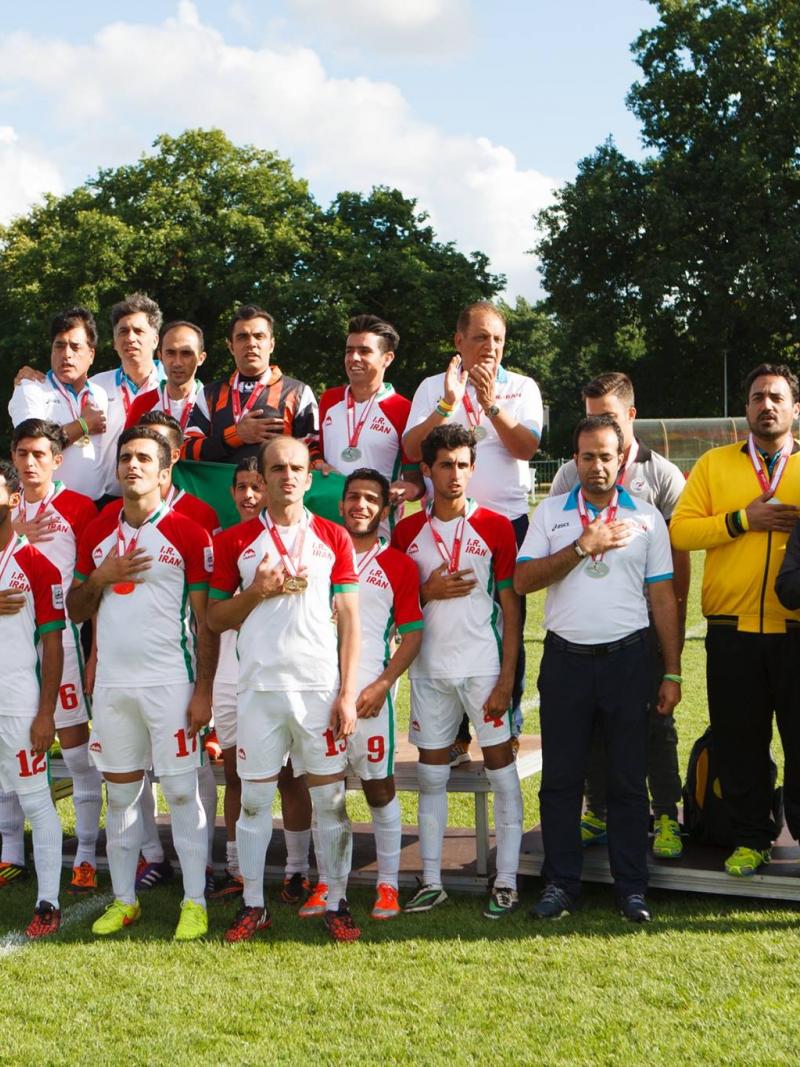 Iranian football 7-a-side team and their staff stand together, put their right hand ver their chests.