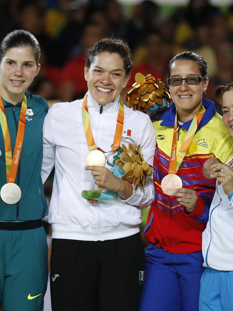 Four women in training suits on a podium, showing medals and mascots