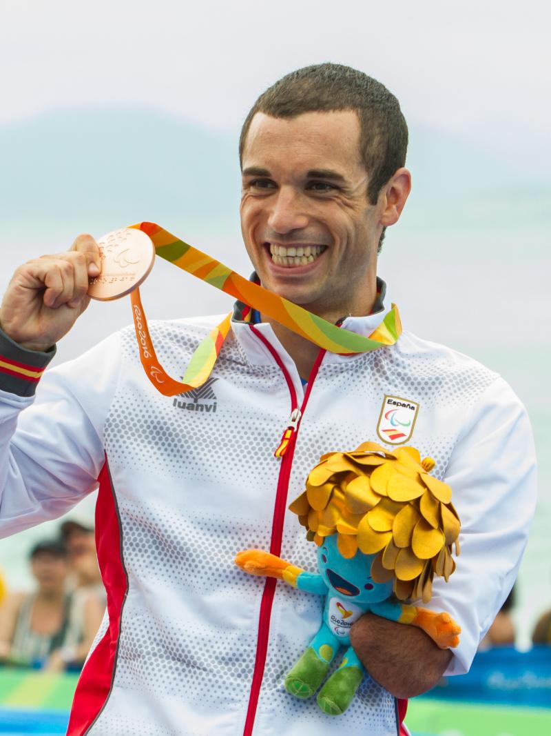 man stands on podium holding up a medal and a mascot