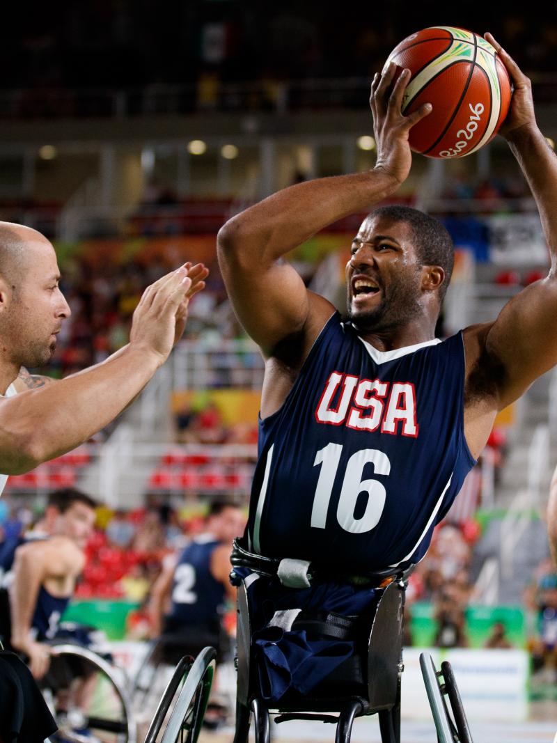 Man in wheelchair holds a basketball in the air away from his defender