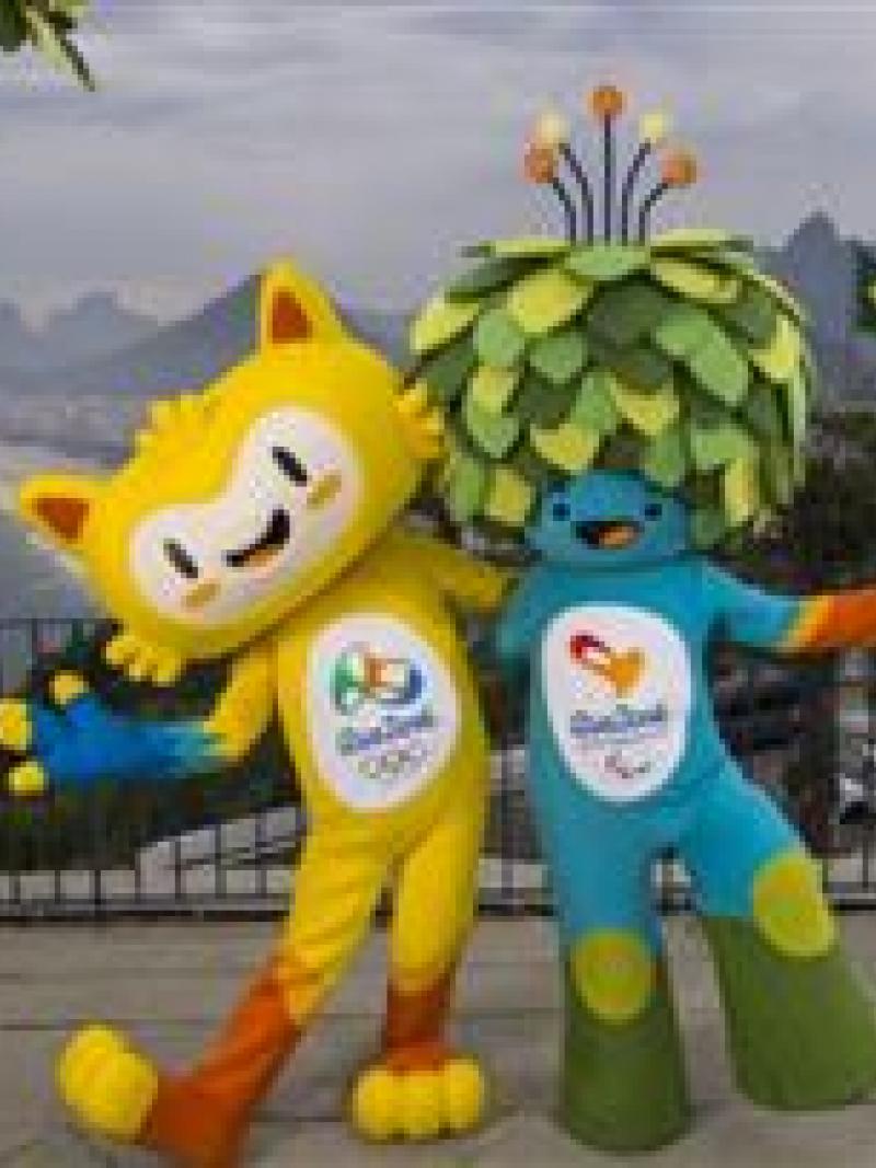 A yellow mascots and blue mascot with green leaves as hair stand on top of Rio's Sugar Loaf mountain.