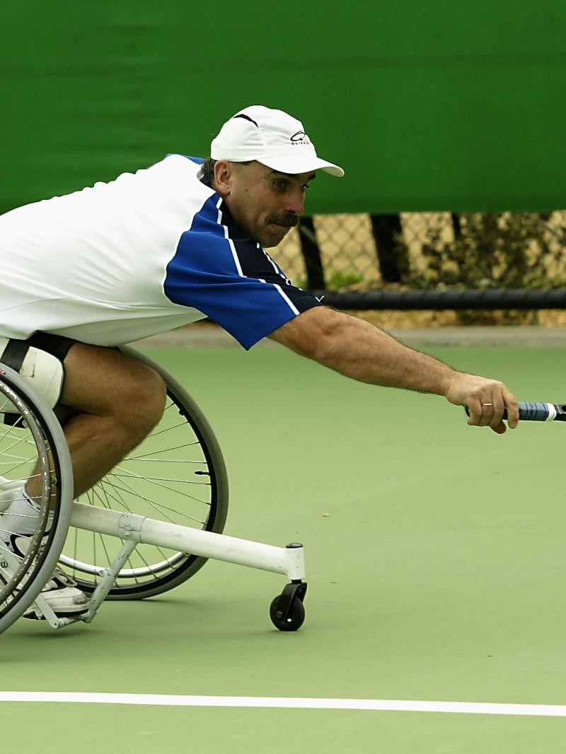 Man in wheelchair reaches to try and hit a tennis ball with his racquet 
