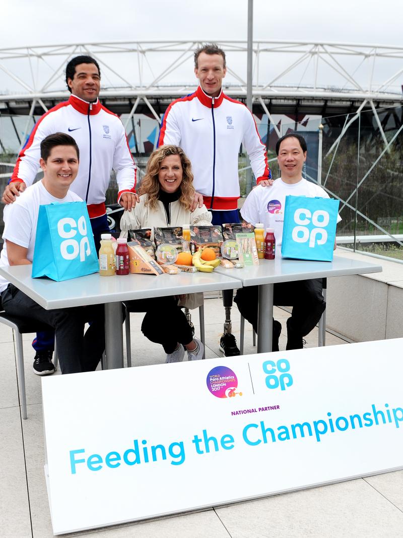 Two men in tracksuits stand behind three people sat at a table.  In the background is an athletics stadium.
