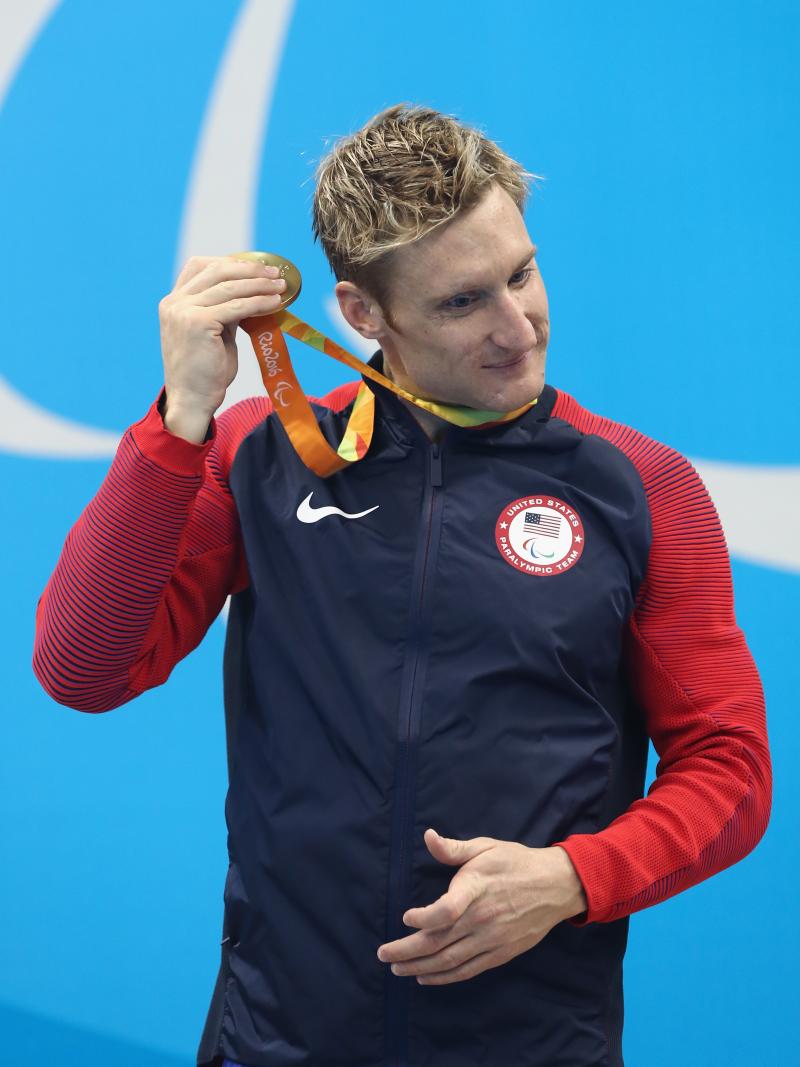 Blind man on podium shakes his gold medal near his ear