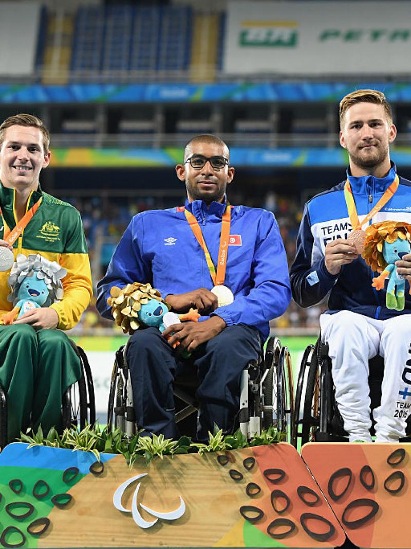 three men in wheelchairs smile holding their medals on the podium