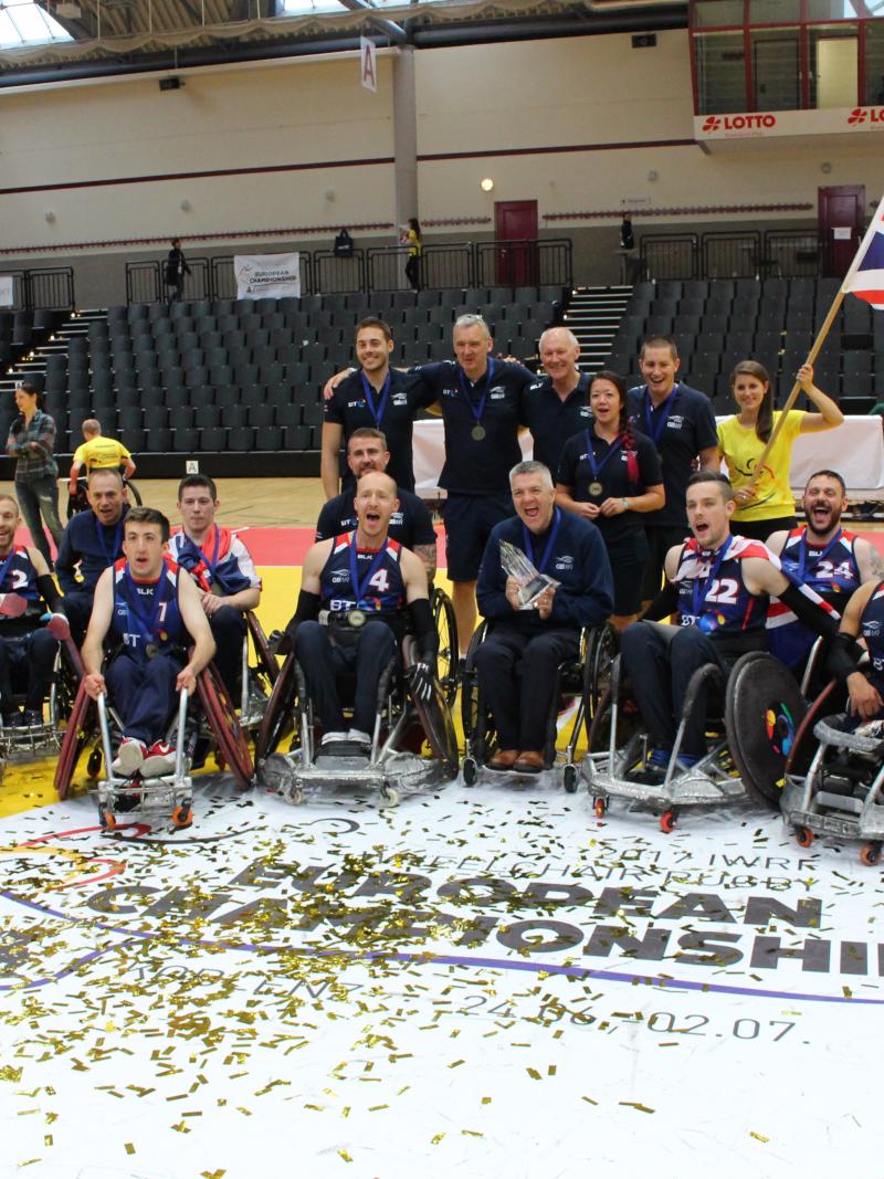 A group of men in wheelchairs surrounded by confetti and ribbons celebrate with the trophy