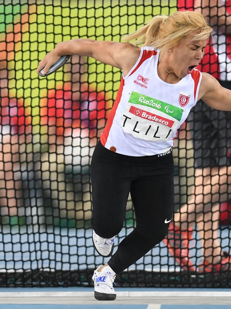 A woman in white vest and black bottoms pulls a face as she gets ready to throw a discus.