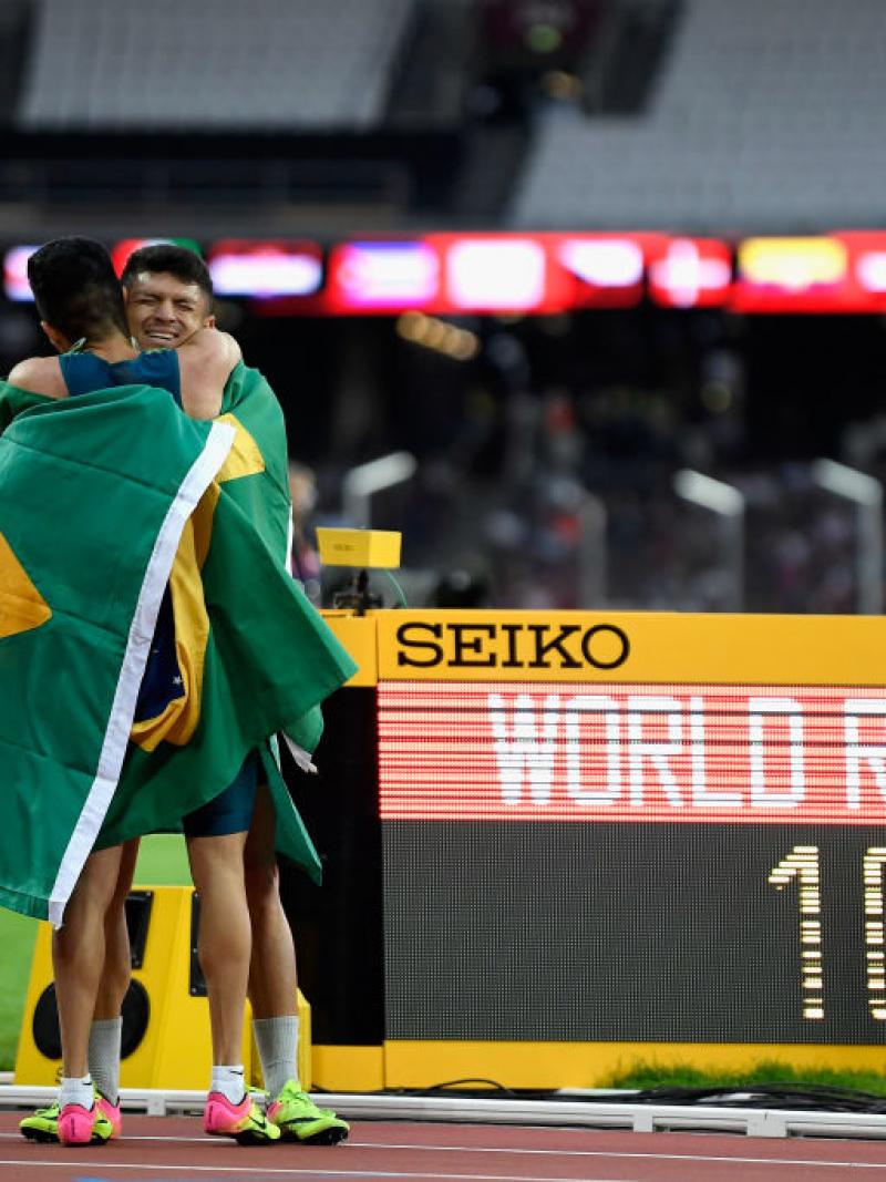 Two athletes hug on the track in front of a bulletin board showing 'WR'
