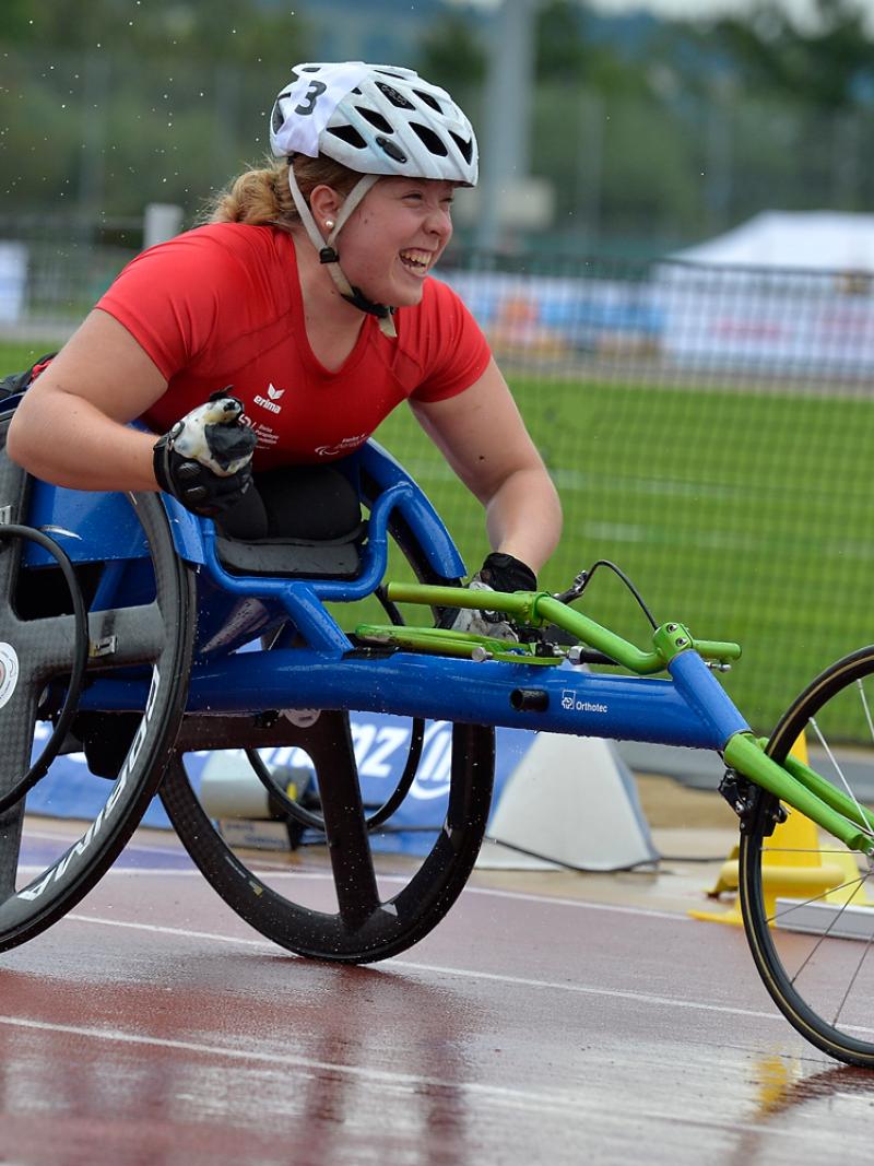 Athlete in a wheelchair celebrates clencing her first with a big smile on a wet track.