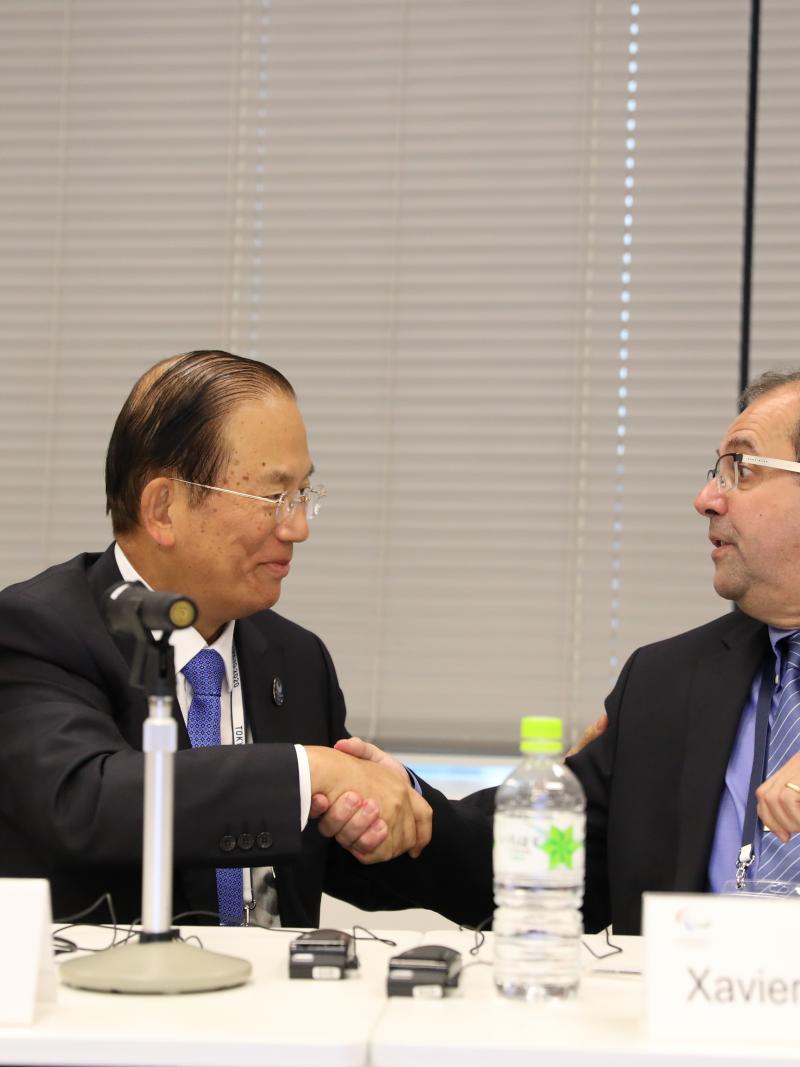 two men shake hands across a table and smile