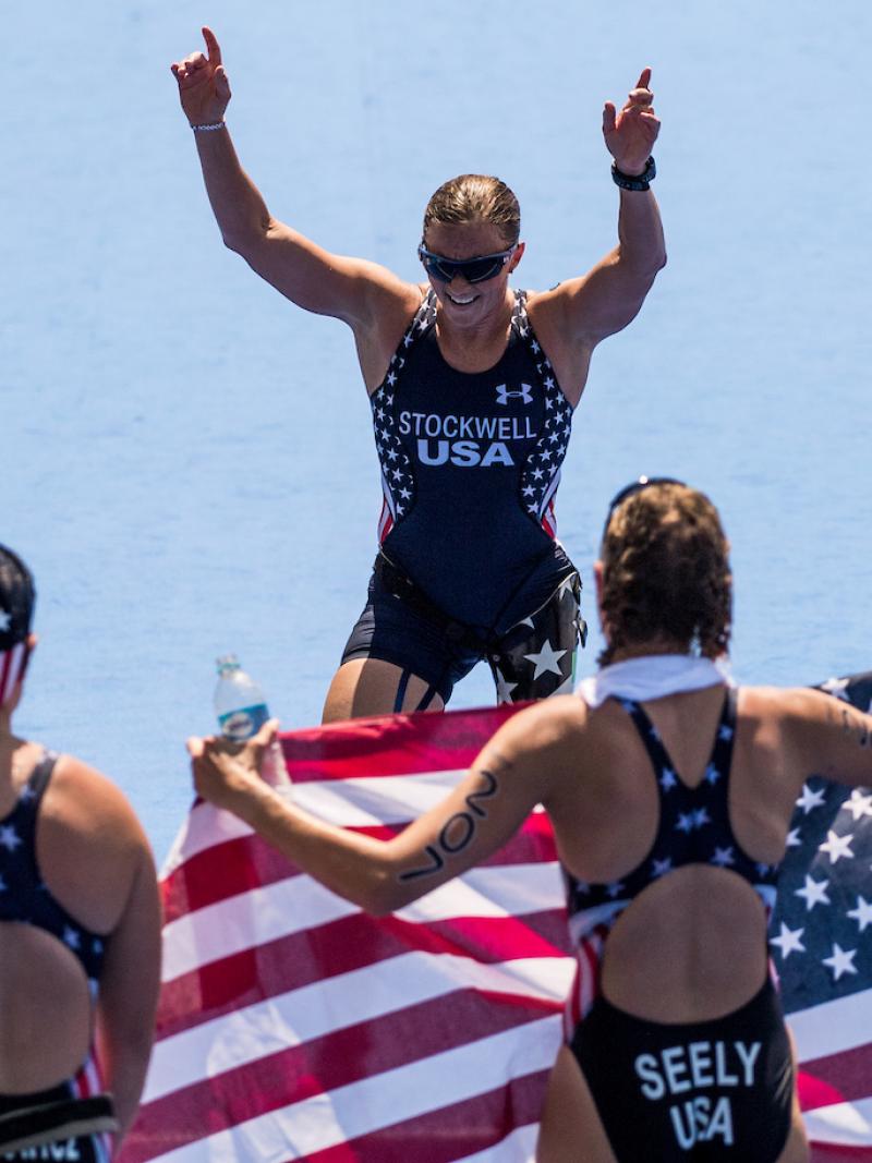 Two women holding the US flag waiting for their teammate to cross the finish line
