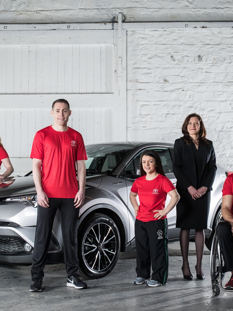 Seven people in front of a car. Six are standing and one is in a wheelchair