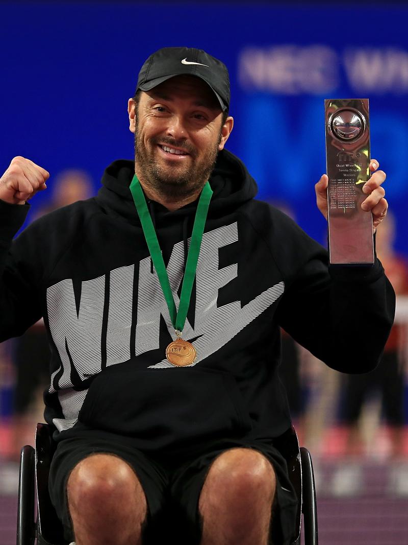 a male wheelchair tennis player holds up a trophy