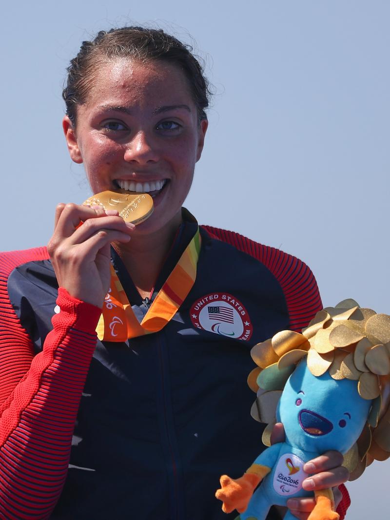 a female Para triathlete stands on the podium and bites her gold medal