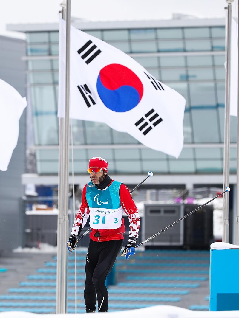 a male Para Nordic skier walks past the Paralympic and South Korean flags