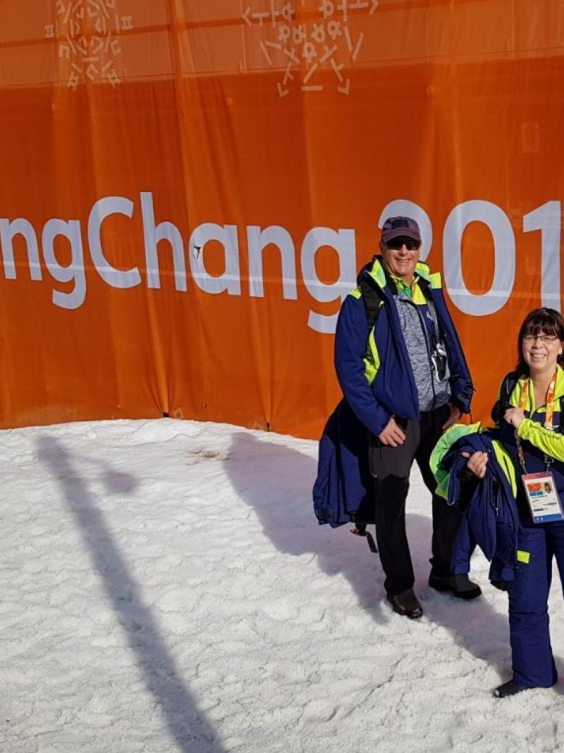 Two winter sport officials pose in front of a PyeongChang 2018 banner