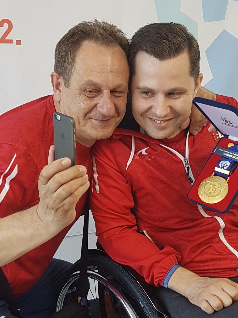 a male Para shooter and his coach hold up his gold medal