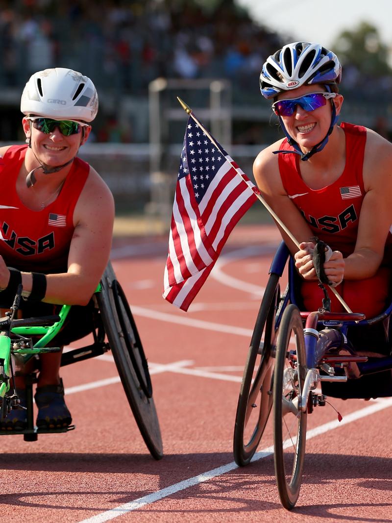 two female wheelchair racers hold up American flags on the track