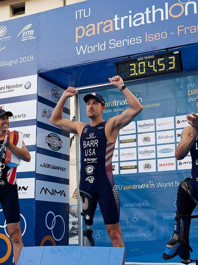 three male Para triathles on a podium with the middle one raising his arms