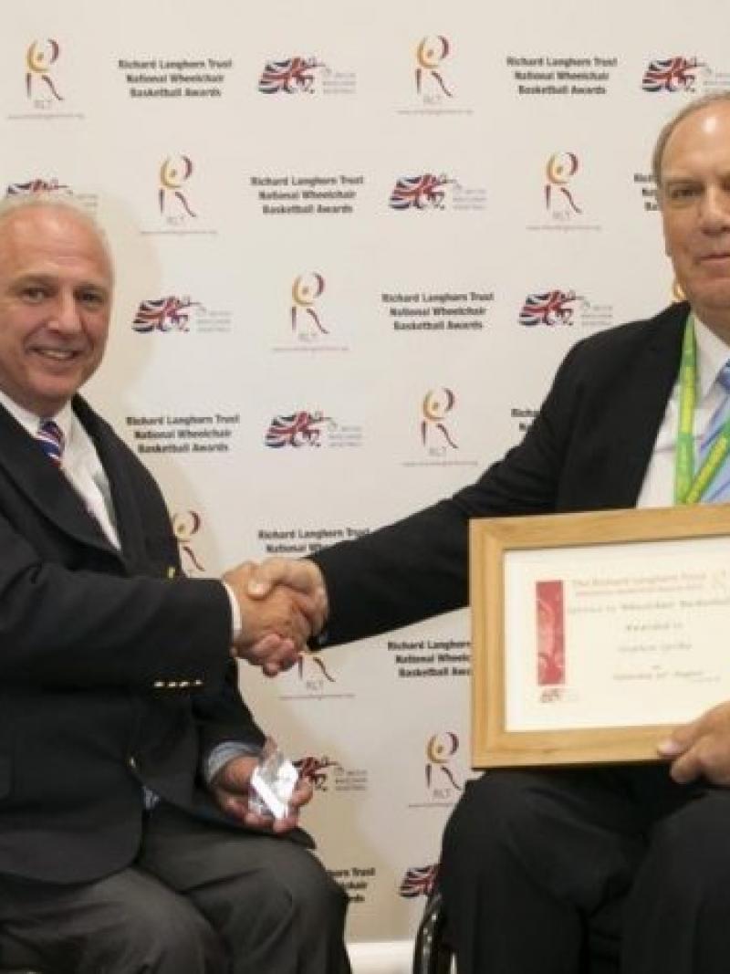 Two men in wheelchair shake hands as one holds a frame