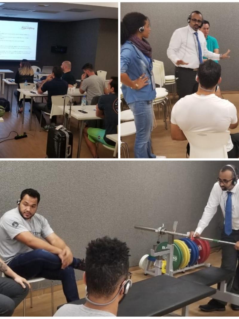 people in a room listening to a man talking and demonstrating a powerlifting bar