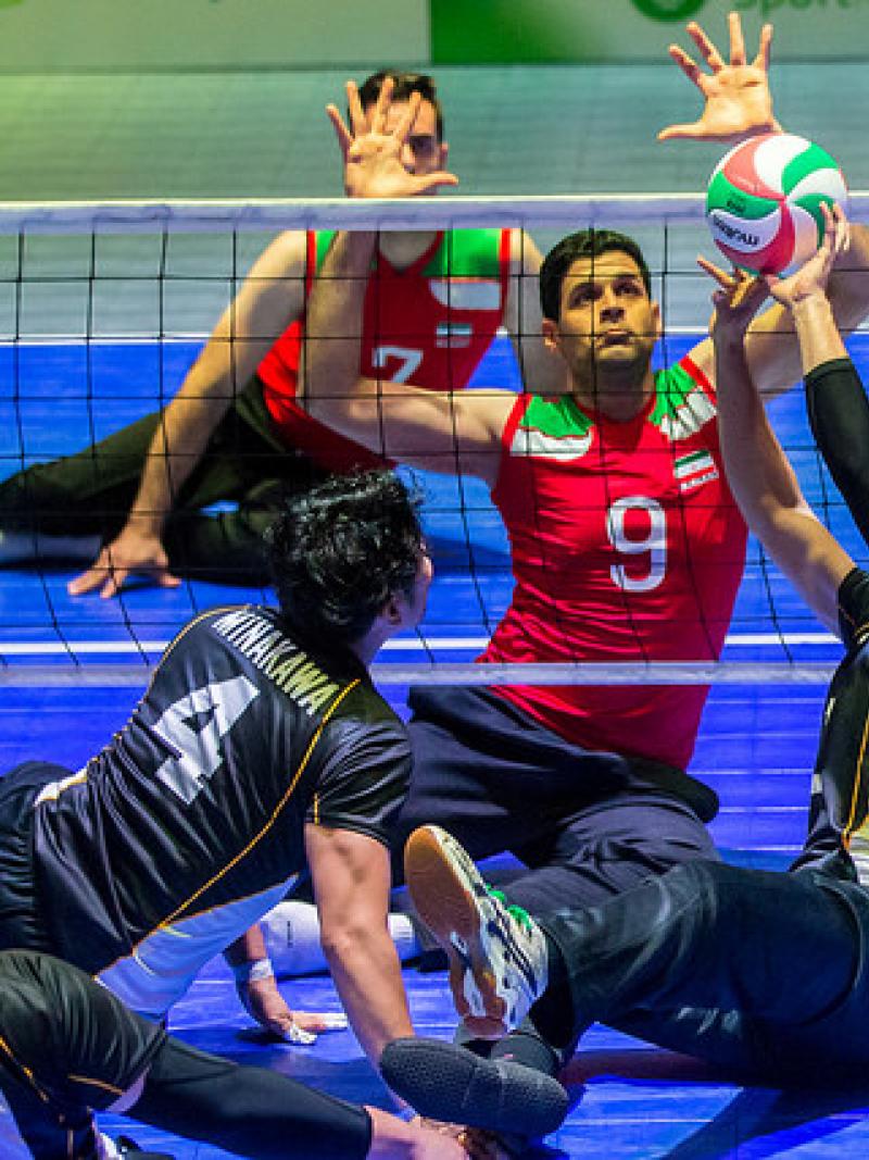 Japanese sitting volleyball player sets the ball while an Iranian athlete prepares to block