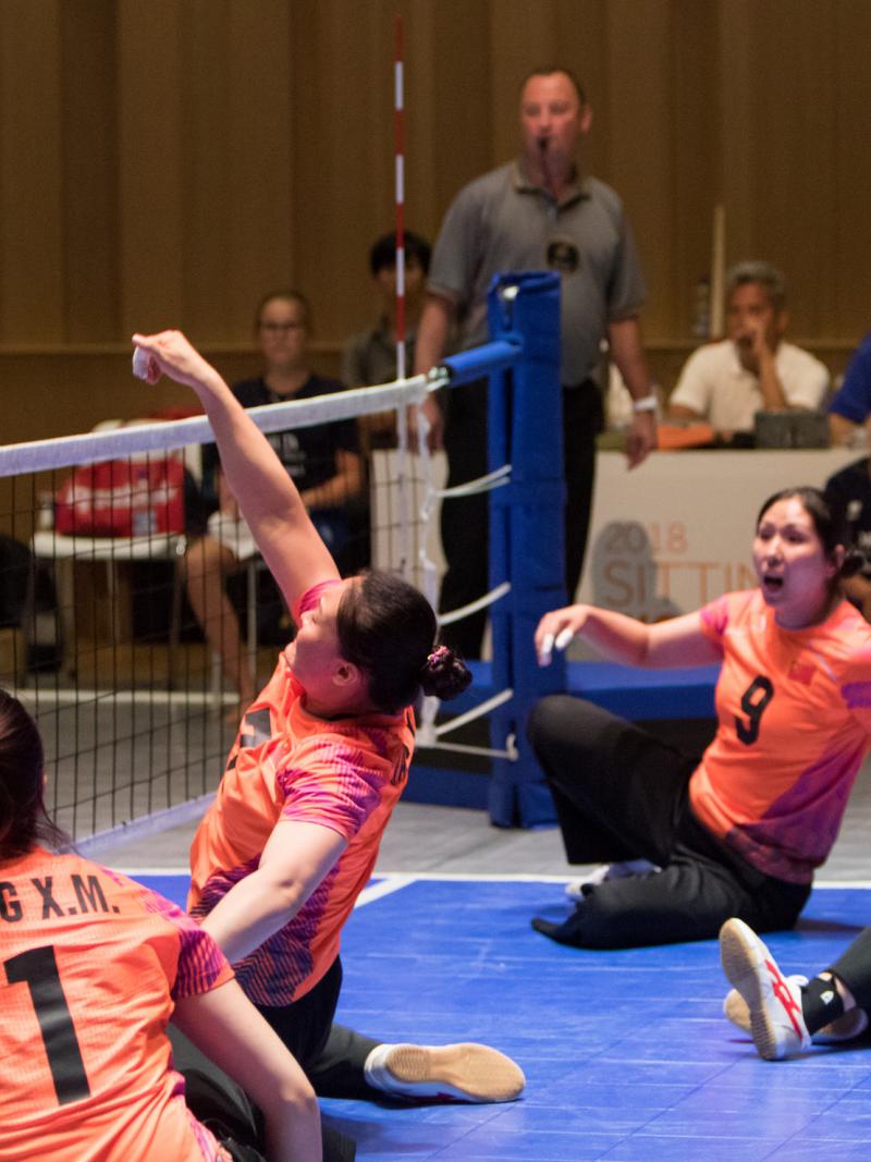 a female Chinese sitting volleyball player punches the ball over the net as her teammates watch