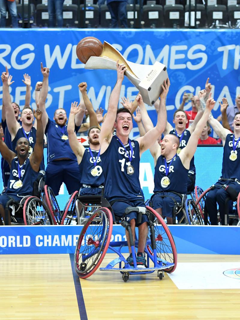 Man in wheelchair hoists basketball trophy with the men's British wheelchair basketball team behind him celerbating 