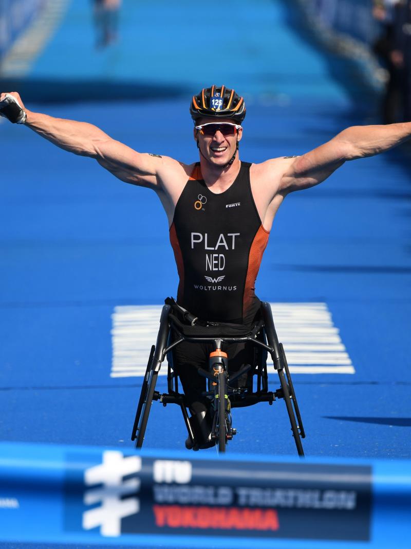 male Para triathlete raises his arms in celebration as he breaks the finish line tape