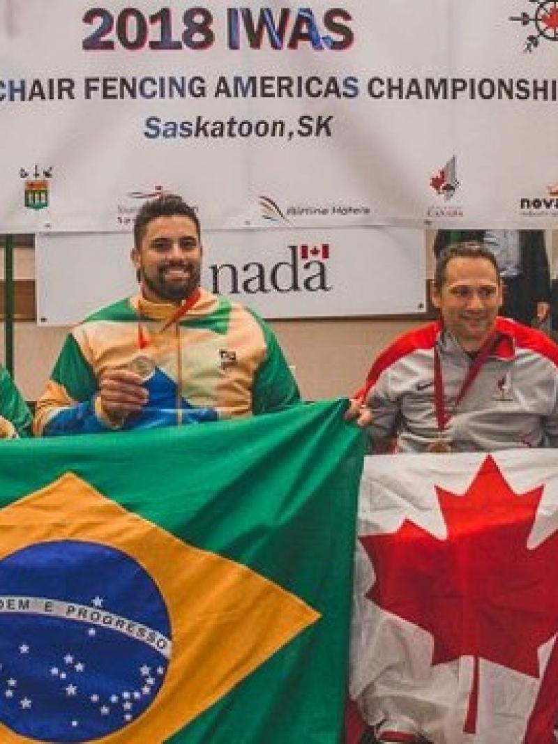 four male wheelchair fencers on the podium including Jovane Guissone holding a Brazilian flag