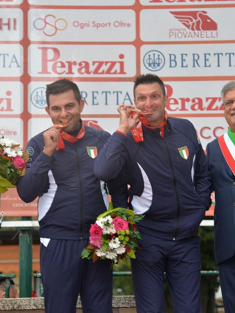 three male Para shooters with Francesco Nespeca in the middle, biting their medals