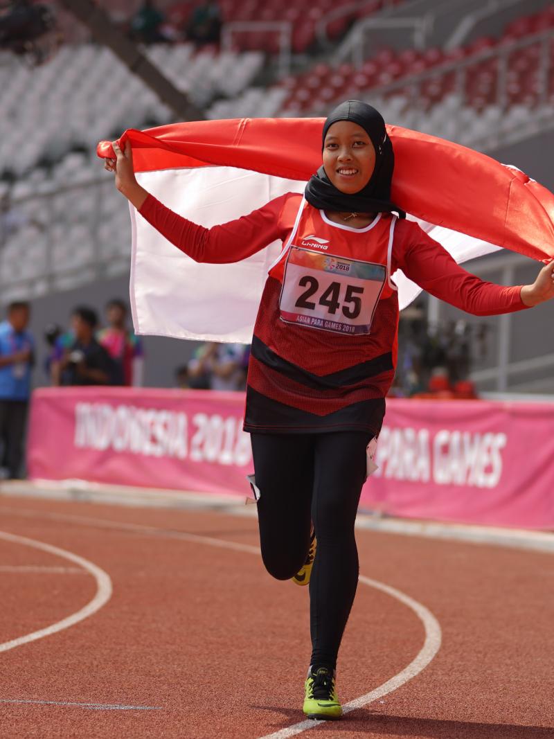 female Para athlete Karisma Tiarani runs down the track holding an Indonesia flag