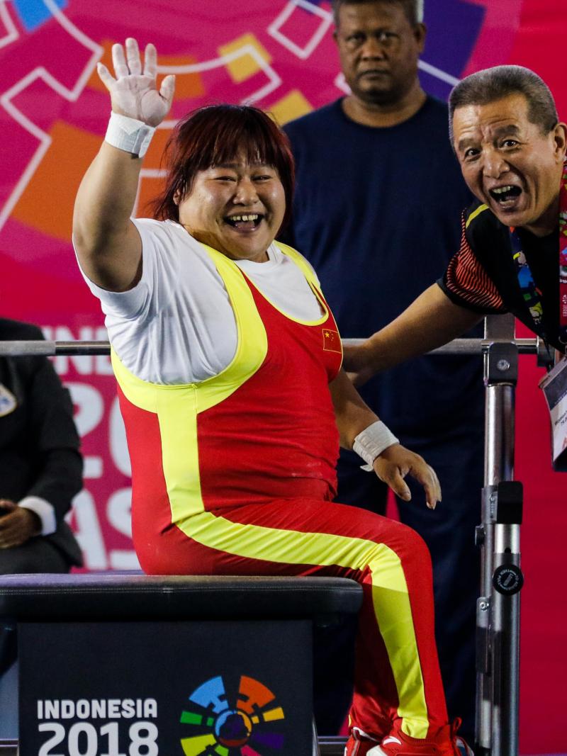 female powerlifter Xu Lili waves and smiles while sitting on the bench