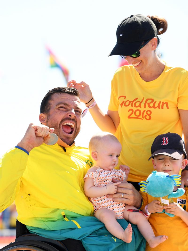 male wheelchair racer Kurt Fearnley holds up his medal alongside his wife and holding his child on his lap