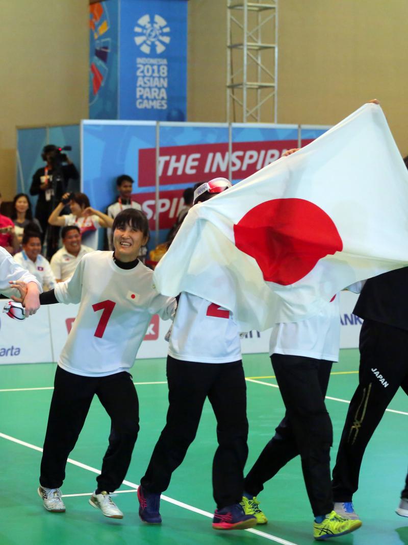 Japanese female goalball players run across the court holding a Japan flag