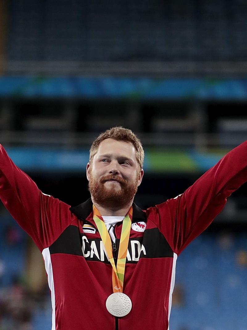 male Para athlete Alister McQueen raises his arms on the podium
