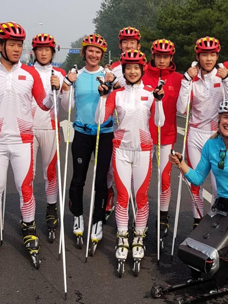 A woman in a sitting rollerski together with a group of ten rollerskiers