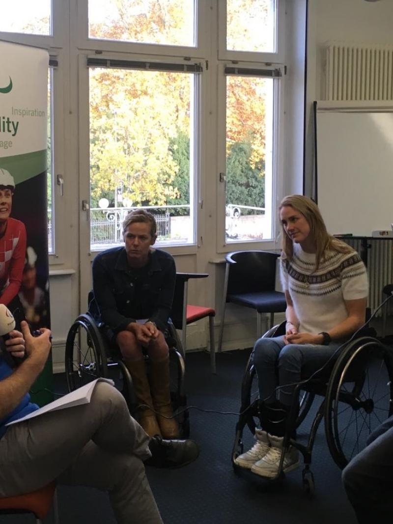 two athletes in wheelchairs watching as a man with a microphone is filmed on a phone