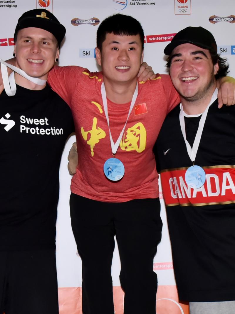 male Para snowboarder Sun Qi stands on the top of the podium with his arms around two other snowboarders