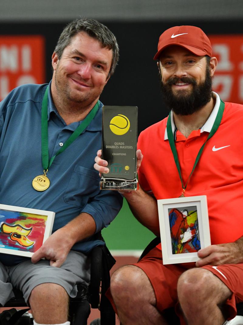 Two men in wheelchair tennis holding a trophy on a tennis court