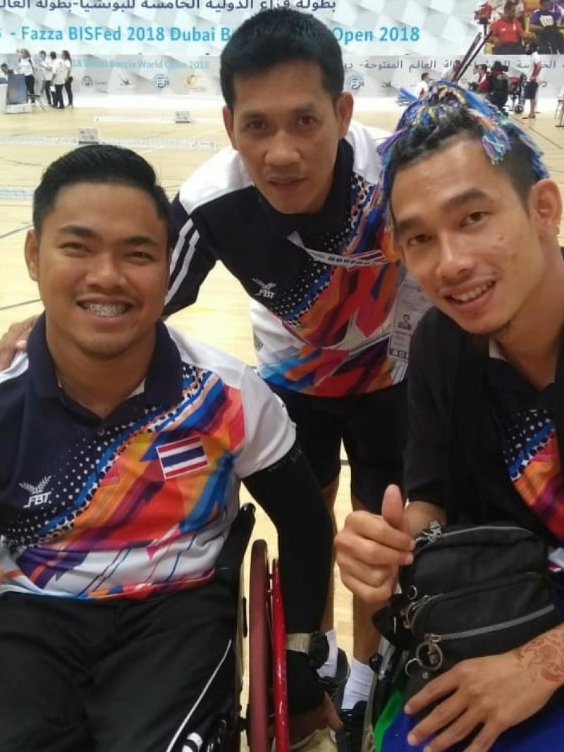 two Thai boccia players and their coach standing behind them and smiling with his arms around their shoulders