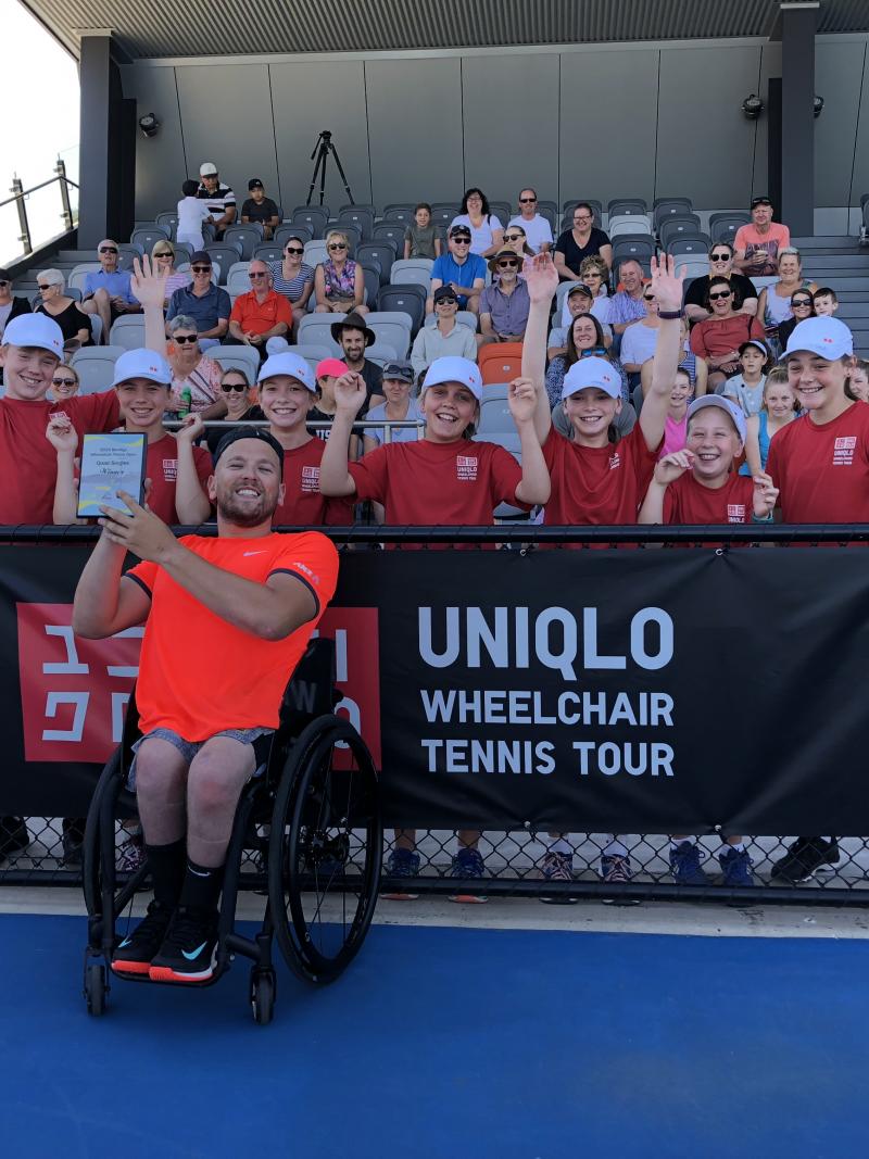 male wheelchair tennis player Dylan Alcott holds up a glass trophy in front of a group of schoolchildren