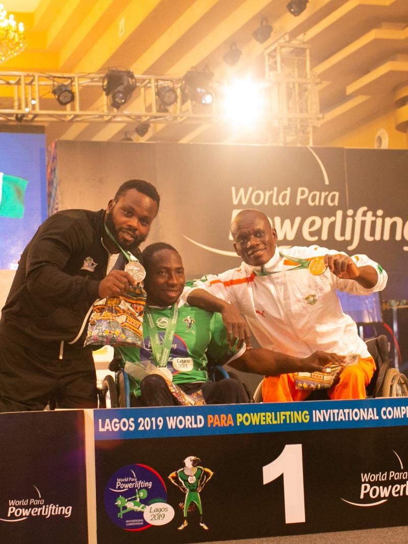 three male powerlifters, with Mufutau Rasaq in the middles, link arms on the podium