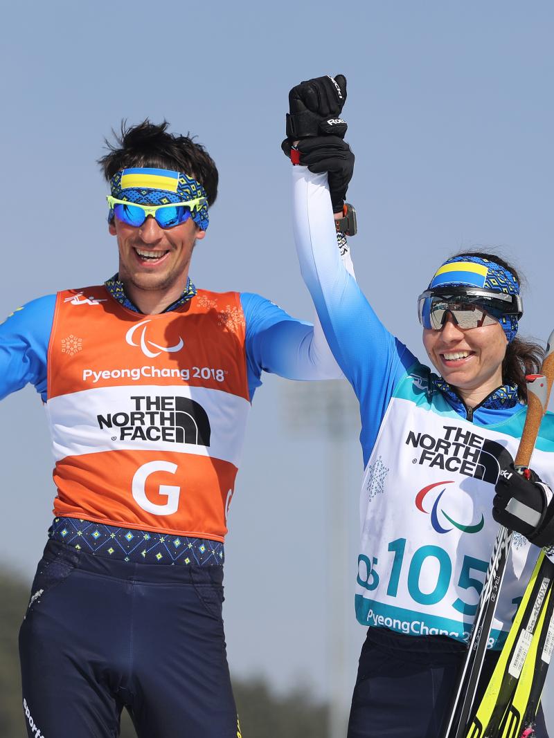 female Para Nordic skier Oksana Shyshkova holds hands with her guide as they raise their arms in victory