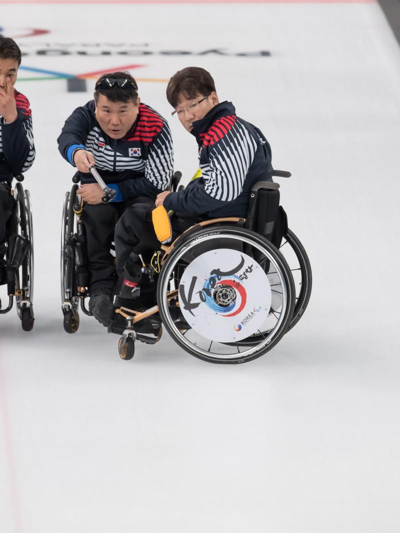 three male wheelchair curlers with Soon-Seok Seo on the right discussing how to play a stone