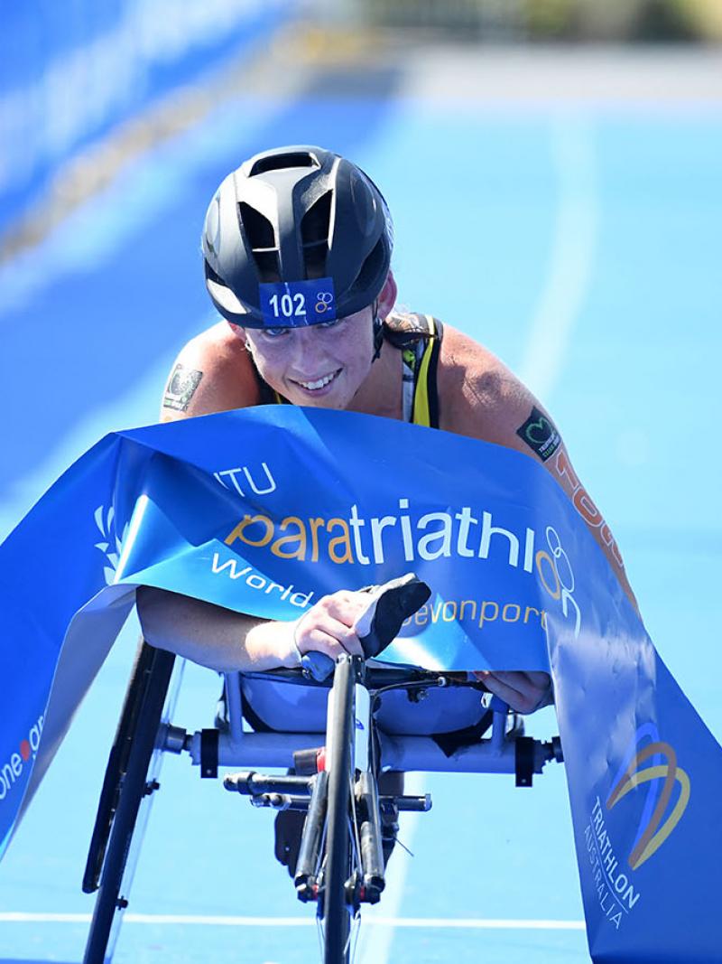 Australian woman in racing wheelchair crosses the finishline tape at a triathlon event