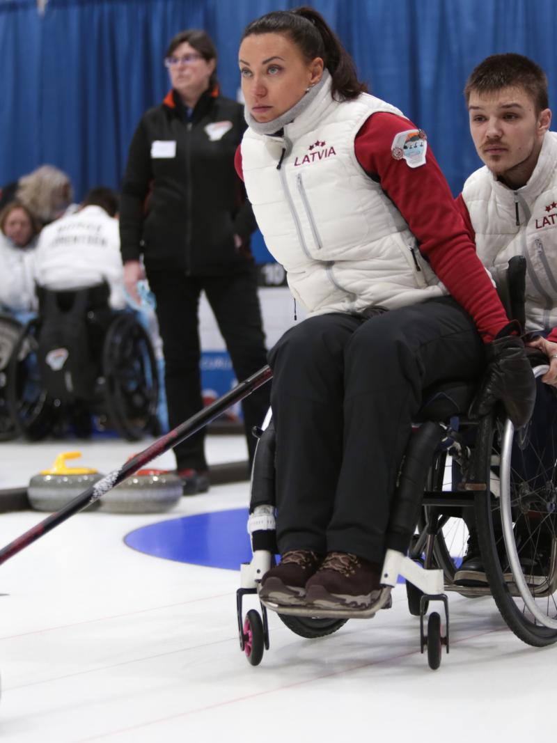 a female wheelchair curler from Latvia plays a stone while another curler holds her wheelchair to steady it