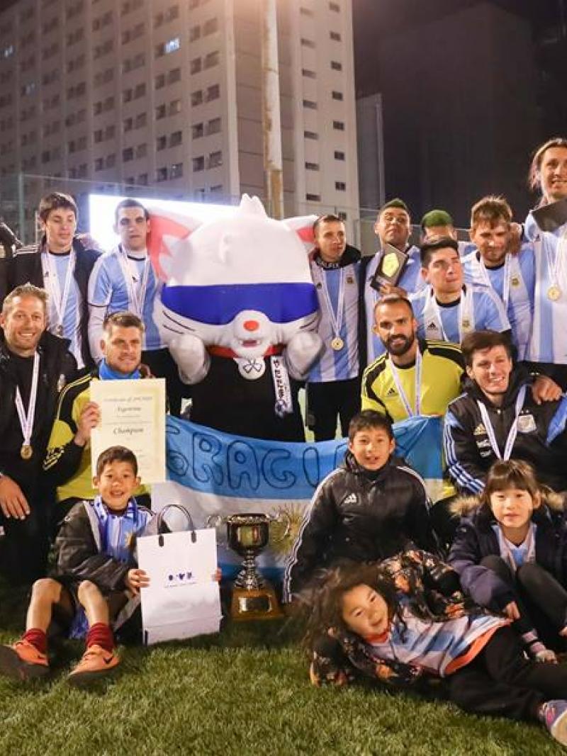 A group of male blind footballers stand with a blindfolded mascot holding a trophy and the Argentinian flag