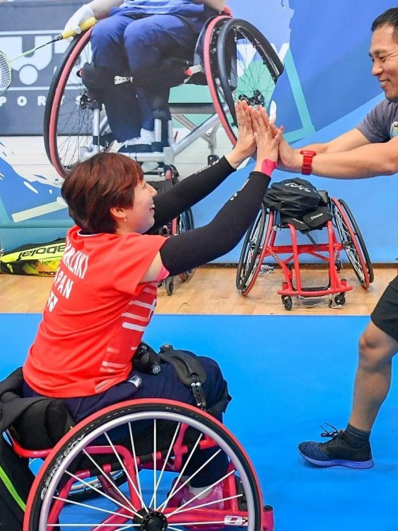 female Para badminton player Yuma Yamazaki sits in a wheelchair and high fives a standing man